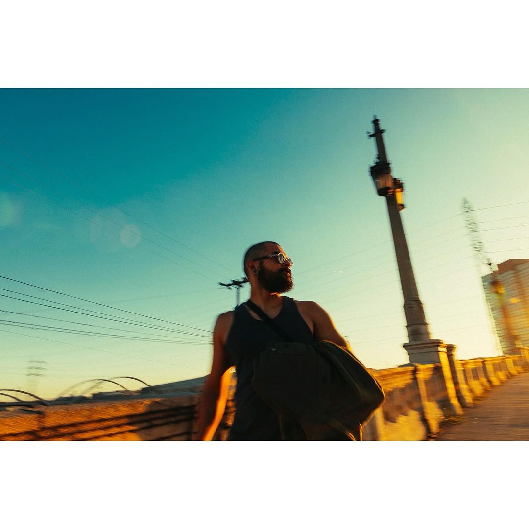Bearded man with glasses and a shoulder bag walks along a bridge with a golden sky and city buildings at dusk or dawn.
