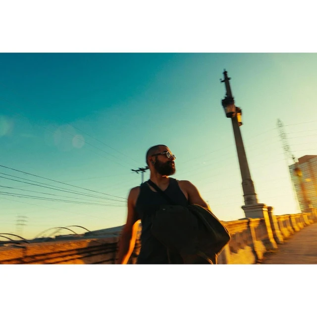 Bearded man with glasses and a shoulder bag walks along a bridge with a golden sky and city buildings at dusk or dawn.