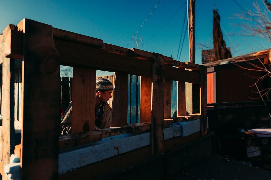 Man in beanie looking through wooden frame structure in an outdoor environment with a vibrant blue sky and surrounding buildings.