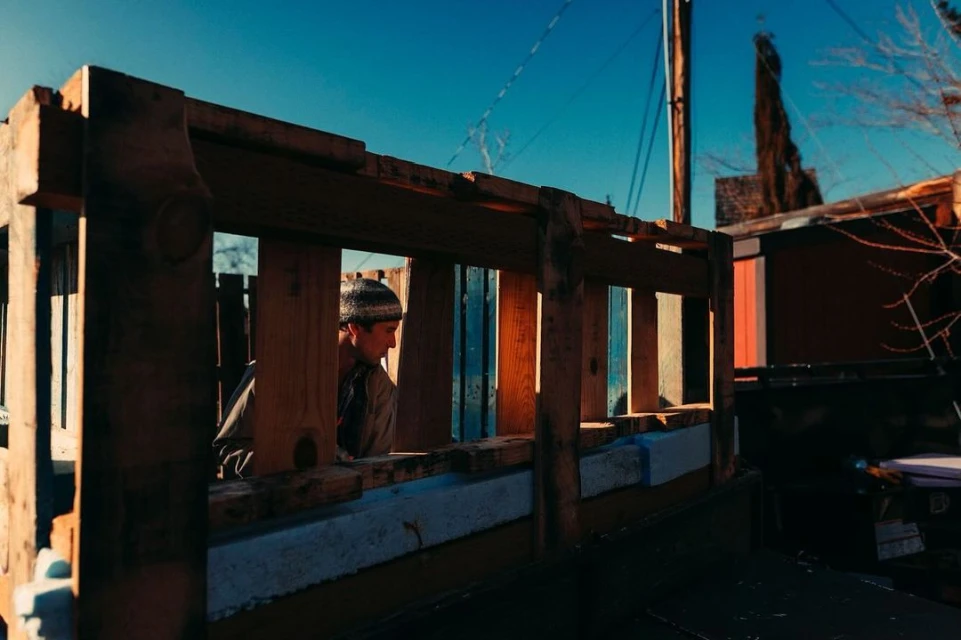 Man in beanie looking through wooden frame structure in an outdoor environment with a vibrant blue sky and surrounding buildings.