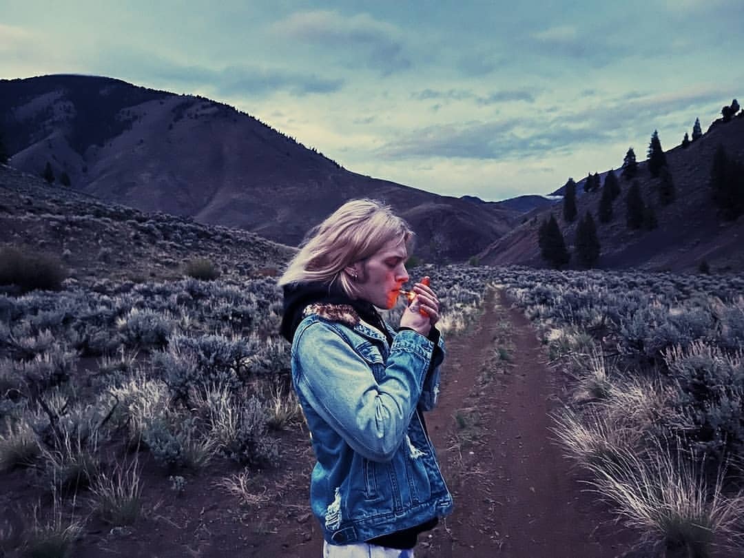 Adult man in denim jacket lights cigarette in outdoor mountain setting with sagebrush under overcast sky. Medium shot.
