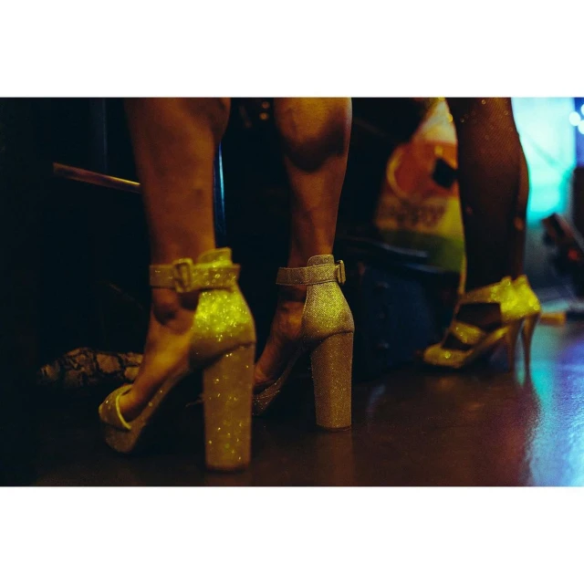 Three figures stand in glittering gold platform high heels, with leg and lower body view. Dark indoor setting with glowing background lights.