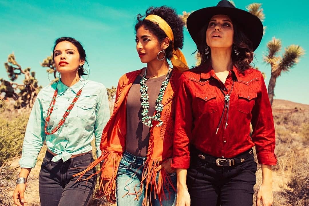 Three stylish women in western wear stand side-by-side in a desert landscape with cacti and a clear blue sky, evoking a vintage fashion vibe.