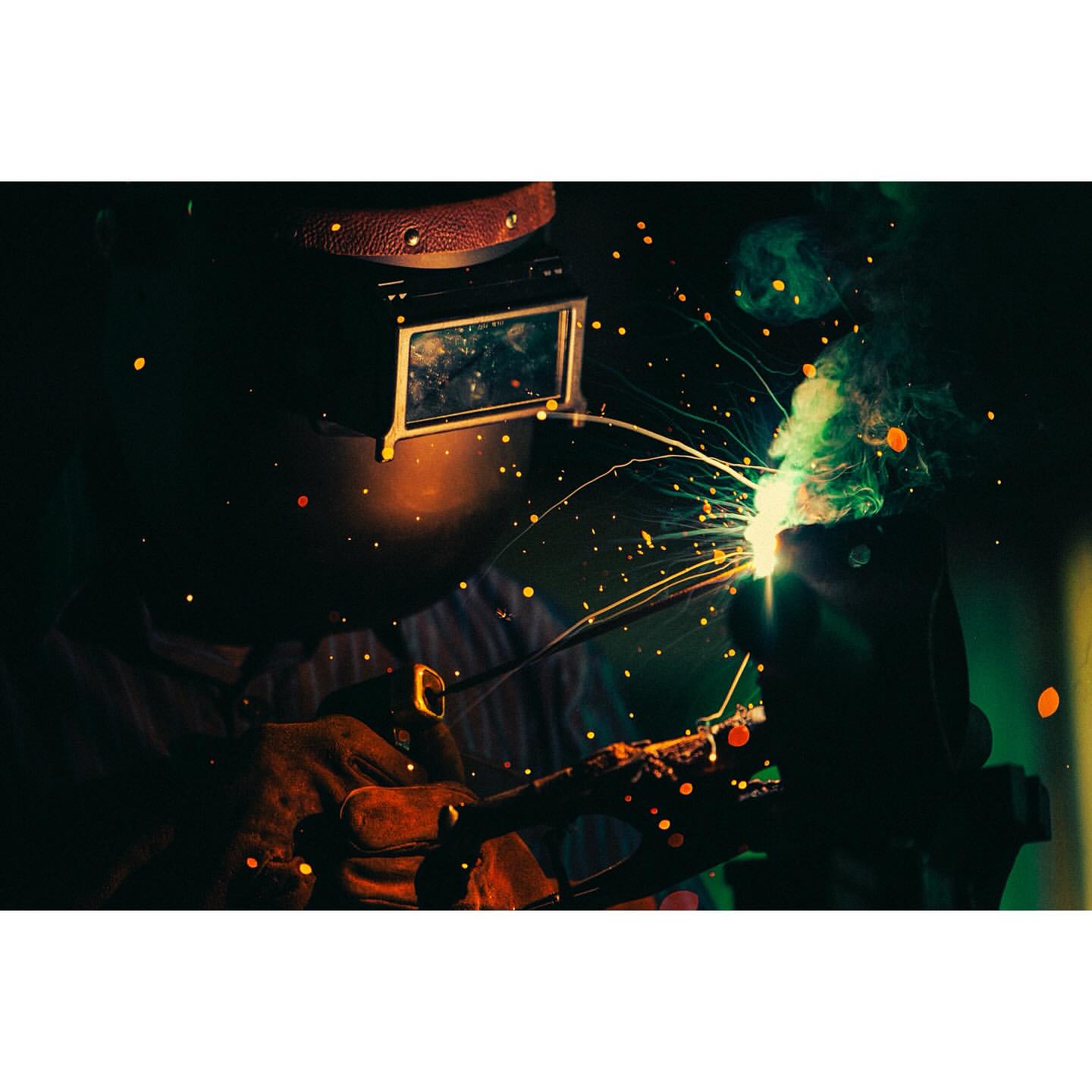 A welder wearing protective gear uses a welding torch with sparks and smoke, set against a dark backdrop with a spotlight effect.