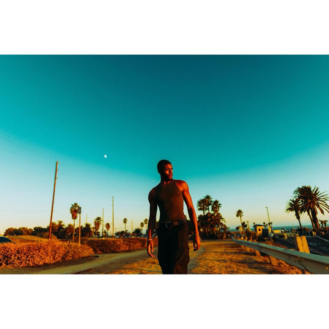Adult male walking along a beachside path lined with palm trees and hedges beneath a bright blue sky and warm golden hour lighting.