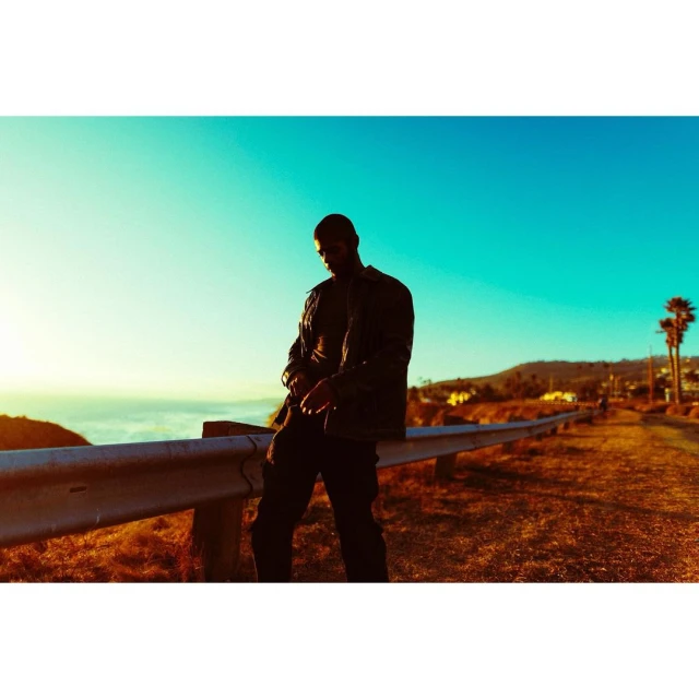 A man in dark clothing stands on a roadside overlooking the ocean under a vibrant blue sky, near a hillside with scattered houses.
