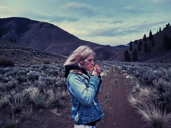 Blonde man wearing a denim jacket lighting a cigarette along a desolate mountain dirt path in San Francisco.