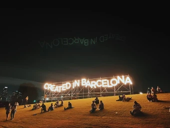 Illuminated neon sign reading CREATED IN BARCELONA on a grassy hill at night with people seated around it