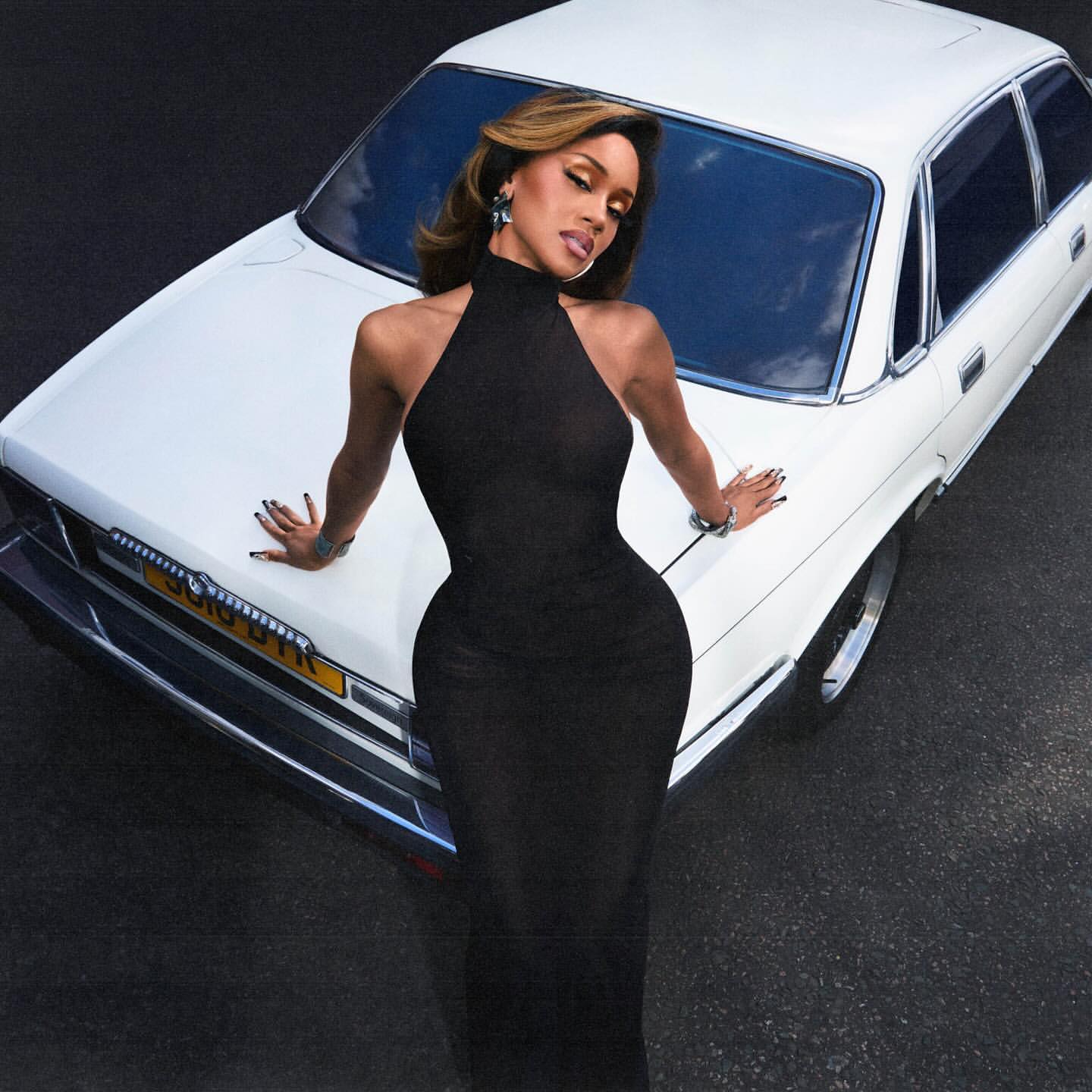 Adult female poses in a black dress next to a white car, creating a high-angle fashion shot with a sleek and stylish aesthetic.