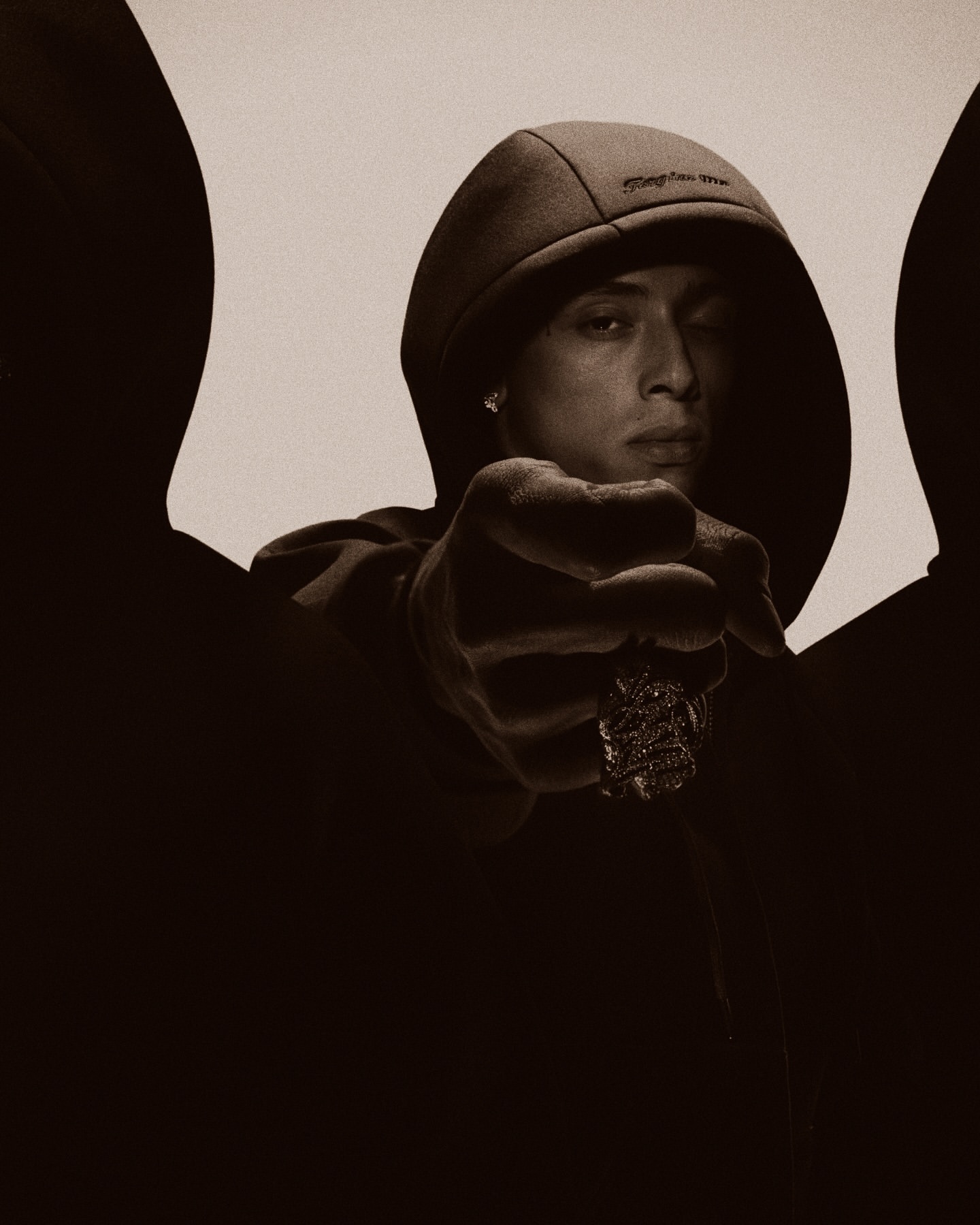 Portrait of a young adult male wearing a hoodie, pointing directly at the camera, and wearing jewelry, in a sepia-toned photo studio setting.