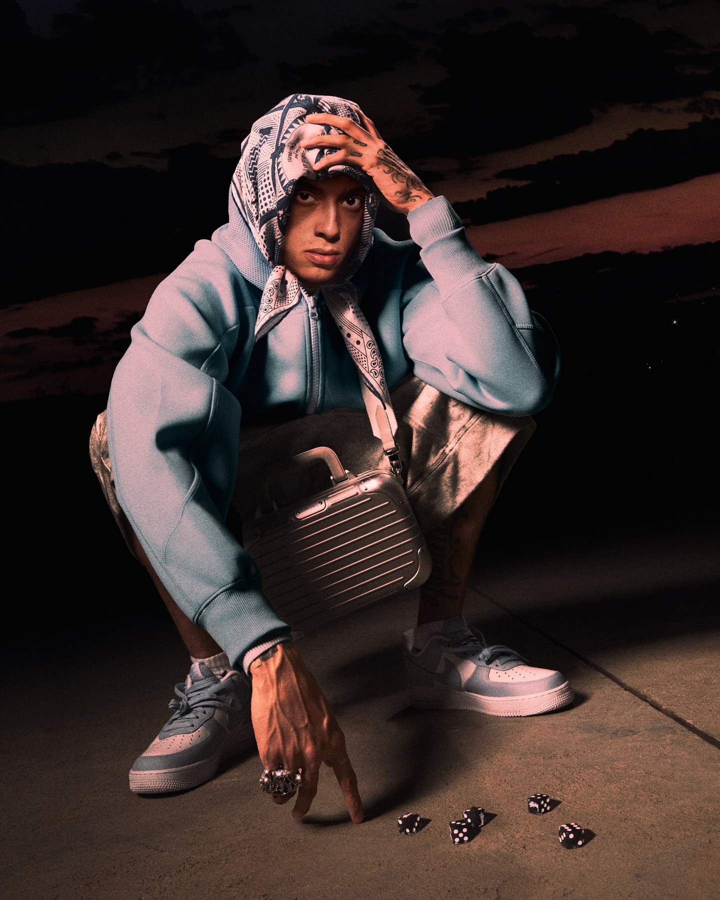 Young adult in hooded jacket and headscarf crouches on pavement with dice at dusk, fashionable streetwear apparel, unique style.