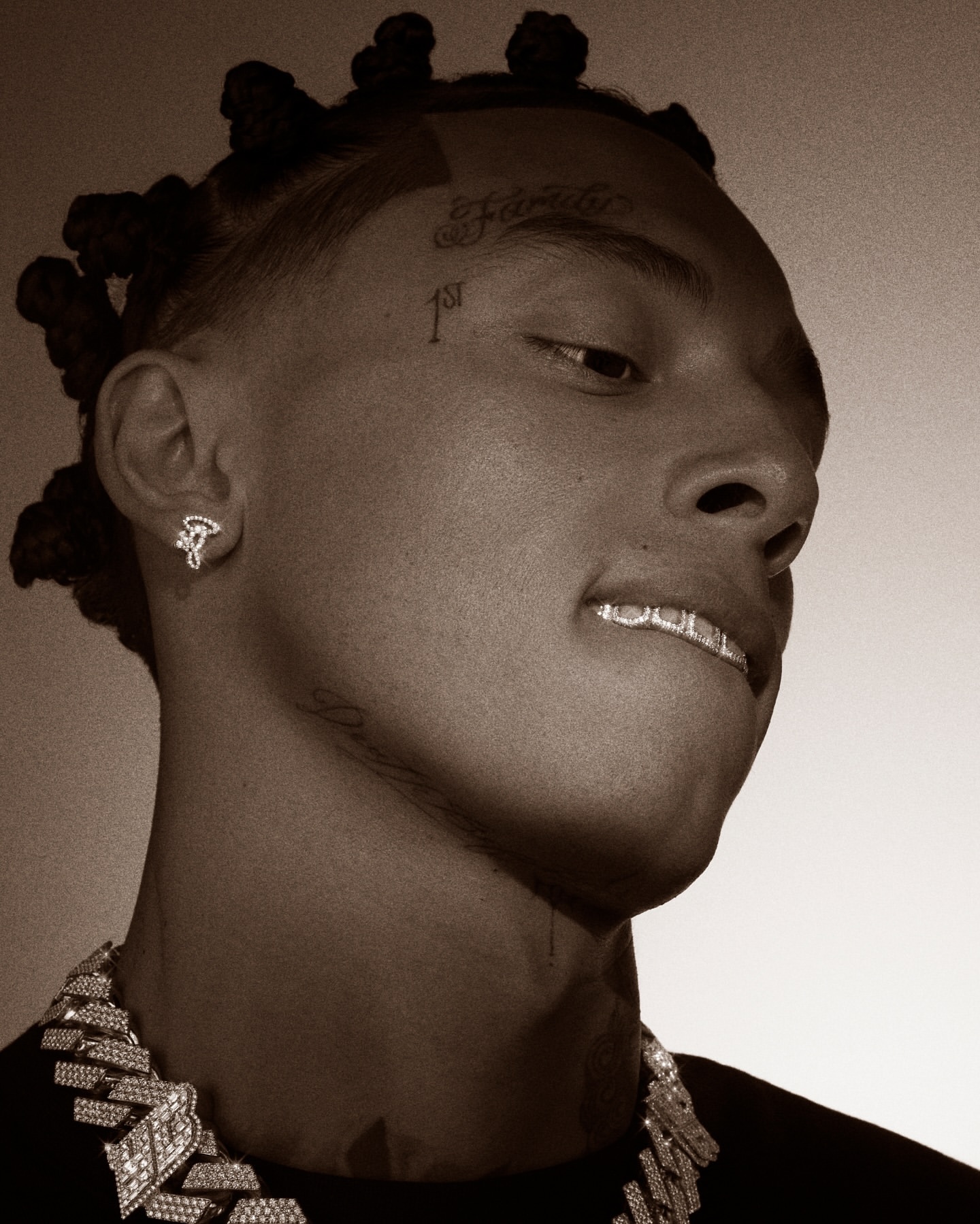Close-up portrait of a young adult male with braided hair and visible face tattoos wearing a diamond chain and grill in a sepia tone.