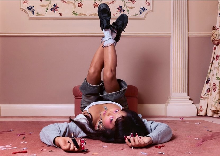 Woman lying on a pink carpet with legs up on ottoman, wearing grey sweatshirt, denim shorts, white socks, and black shoes indoors.