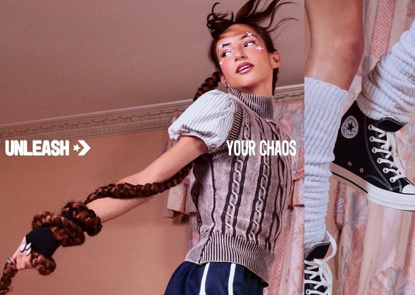 A model poses indoors wearing a cable-knit vest, striped shirt, and sneakers, with makeup accents and a long braid, in a promotional fashion shot.