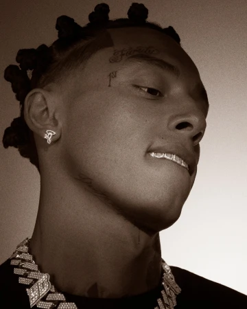 Close-up portrait of a young man with face tattoos, diamond grillz, and braided hair tied in small knots against a neutral background.