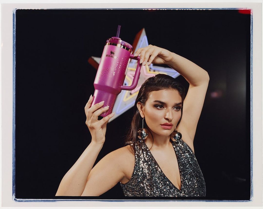 Woman poses in a sequin dress, holding a pink Stanley tumbler with a straw against a black background with a neon star.