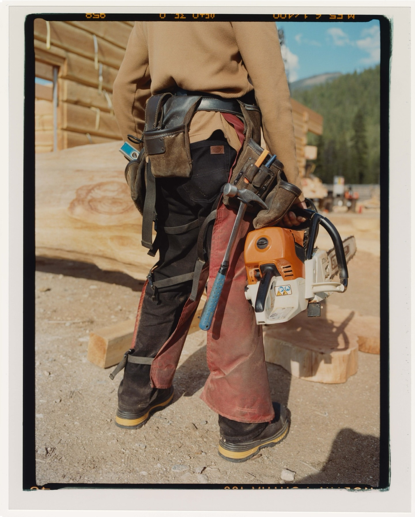 Construction worker wearing tool belt carrying a chainsaw on a dirt construction site with a partially built wood cabin in the background.