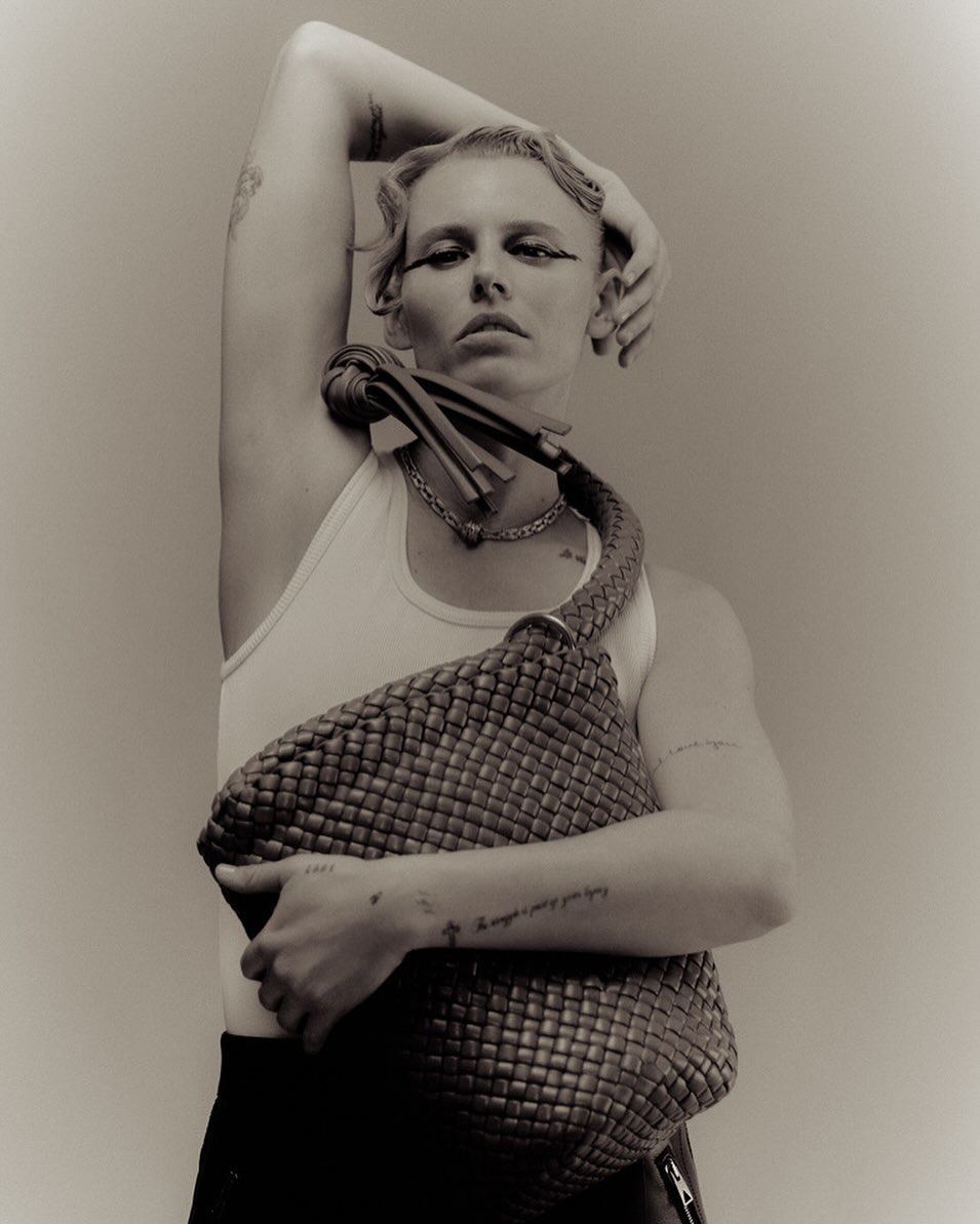 A woman poses indoors, showcasing a Bottega Veneta woven leather handbag. The image features a sepia tone, capturing fashion and style.
