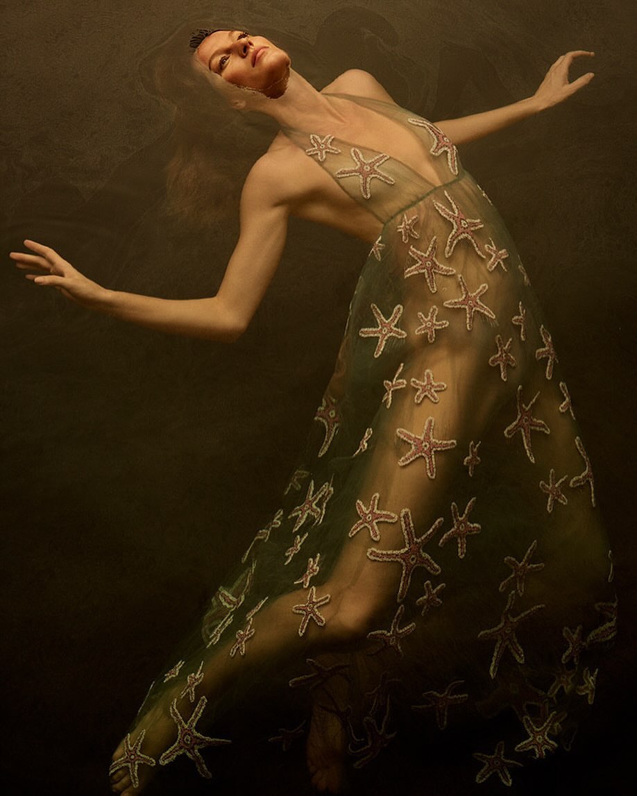 Adult female model in a translucent green dress with starfish appliqués is underwater in a dark setting, artistic fashion photography.