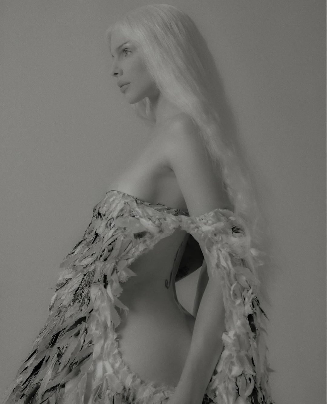 Elegant woman in a high-fashion sculptural dress made of feathers poses in profile against a neutral background, showcasing textures and design.