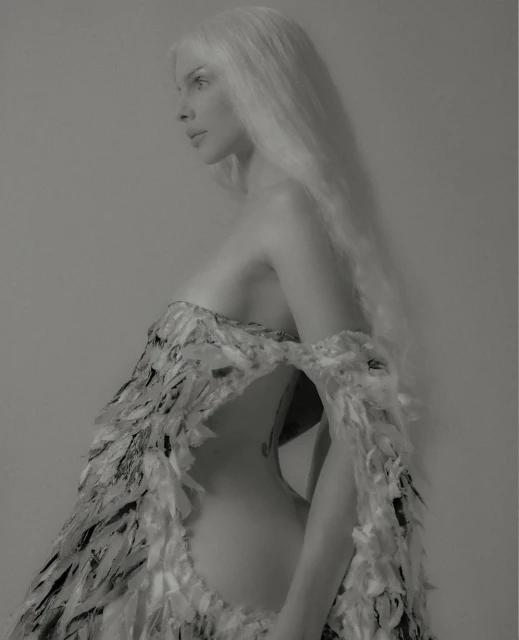 Elegant woman in a high-fashion sculptural dress made of feathers poses in profile against a neutral background, showcasing textures and design.