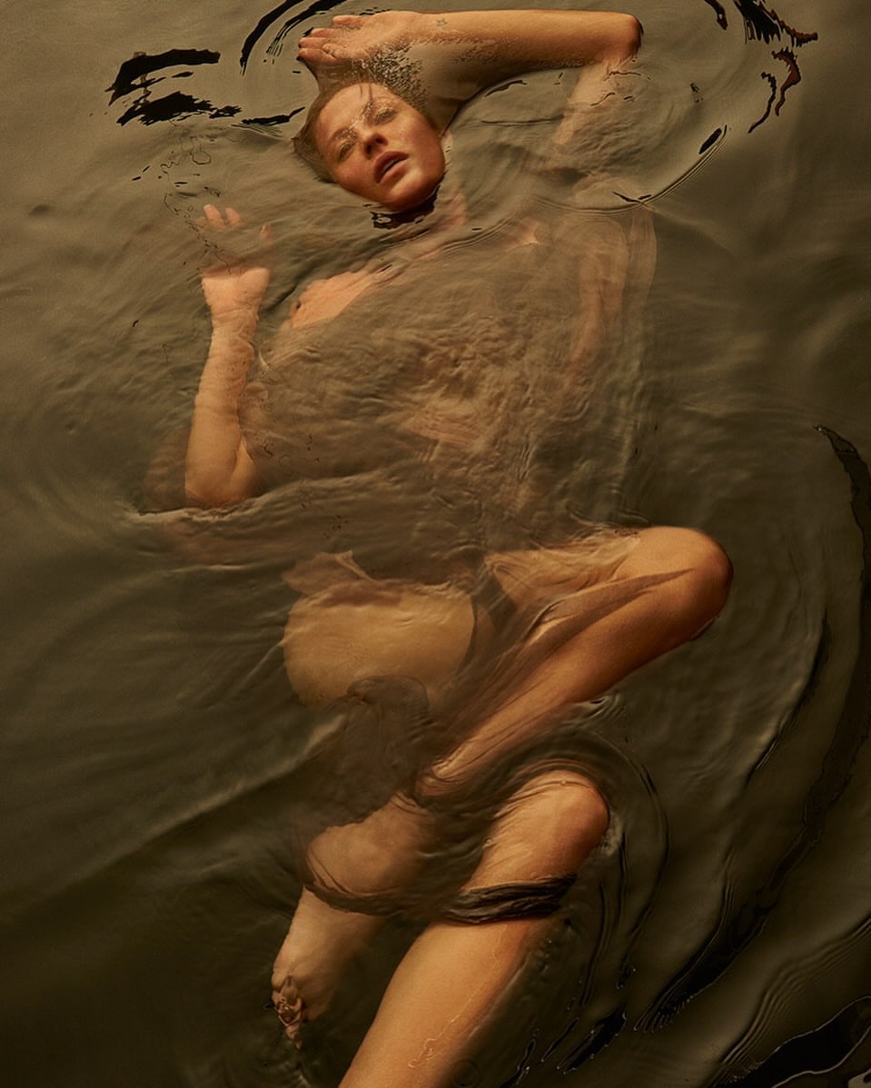 Woman submerged in water with arms raised, creating ripples and reflections, viewed from above in an ethereal, artistic composition.