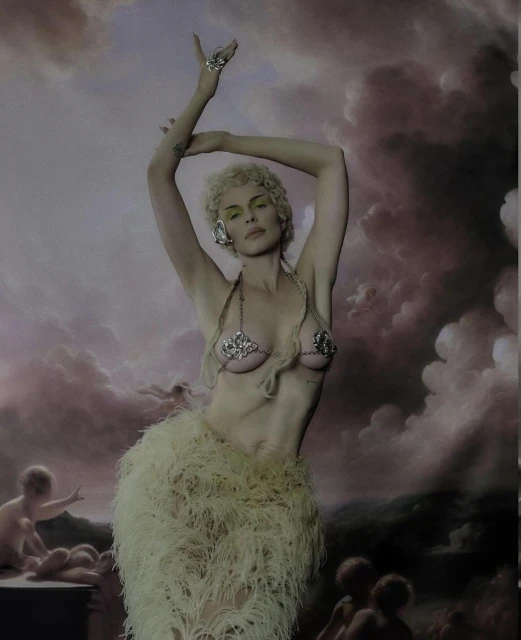 Woman in feathered skirt and jeweled bikini top poses against a painted background.  She has blonde hair and wears ornate jewelry.