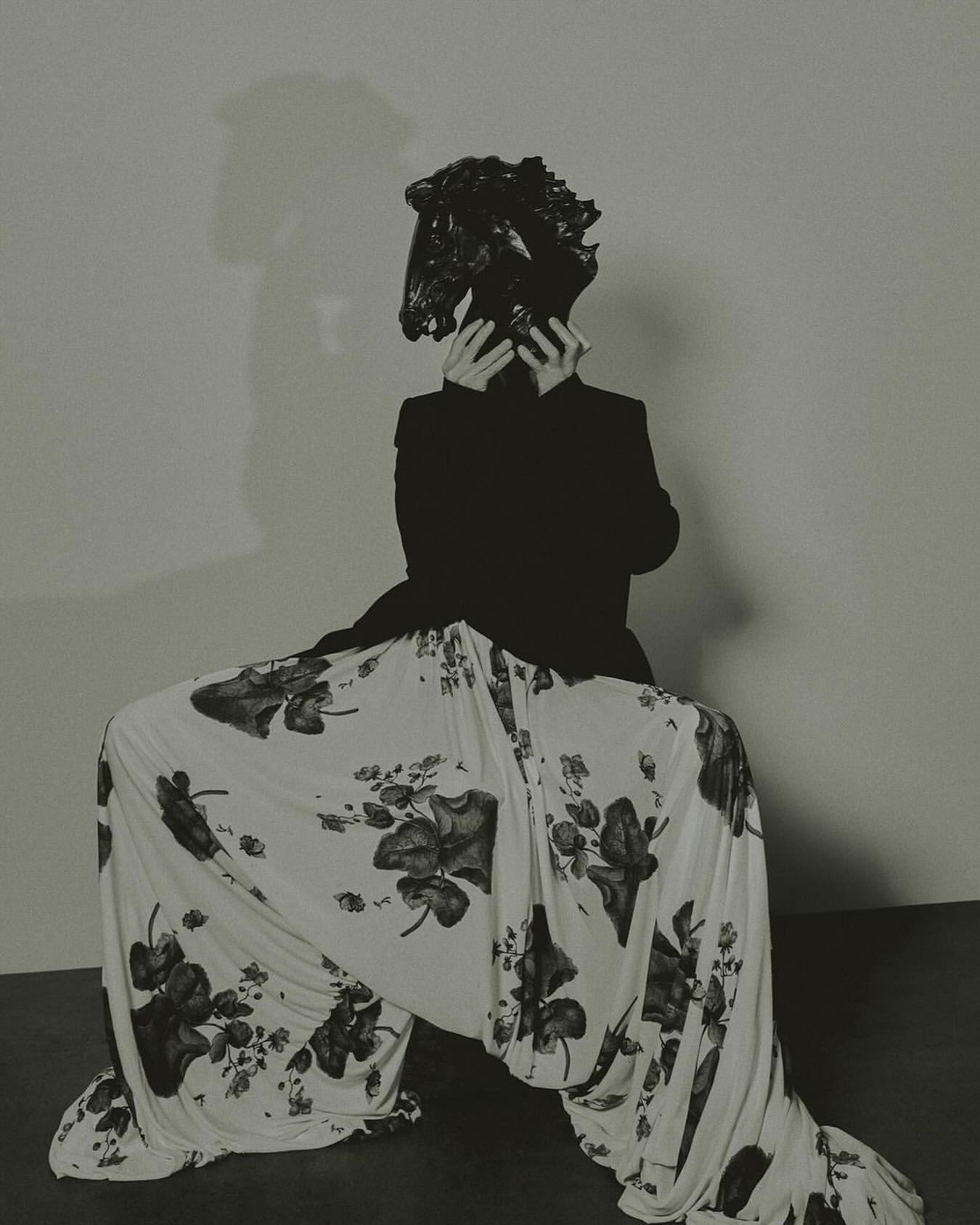 A person in a black blazer and floral pants sits in a squatting position with their face obscured by a black, crumpled plastic bag.