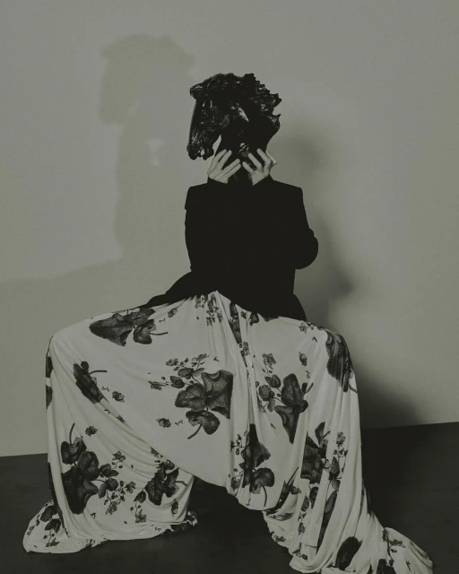 A person in a black blazer and floral pants sits in a squatting position with their face obscured by a black, crumpled plastic bag.