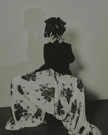A person in a black blazer and floral pants sits in a squatting position with their face obscured by a black, crumpled plastic bag.