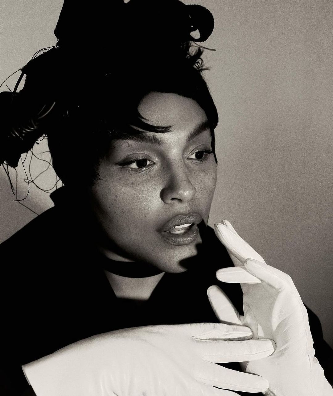 Monochrome portrait of a woman with a head covering and makeup wearing gloves, against a neutral backdrop in a studio setting.