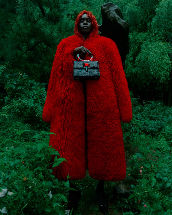 Fashion shot featuring a Black woman in a red coat, holding a black purse, standing in a vibrant green outdoor setting with lush foliage.