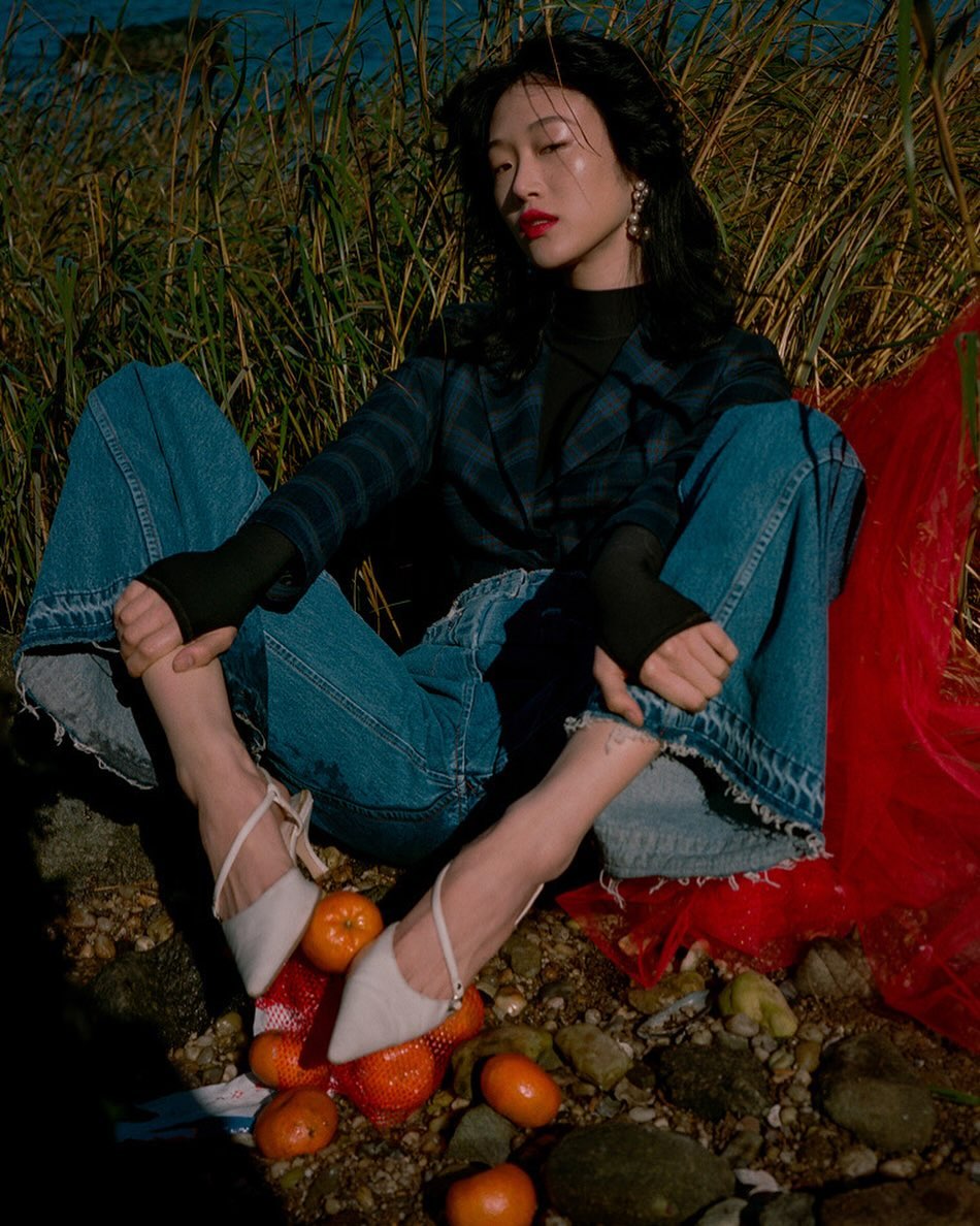 Stylish Asian woman in jeans and blazer sits on rocky ground with oranges, posing in an outdoor fashion shot with natural lighting.