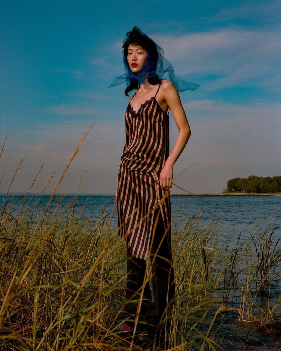 A fashionable Asian woman poses outdoors in a striped dress and sheer blue scarf, amidst tall grass and a seascape backdrop.