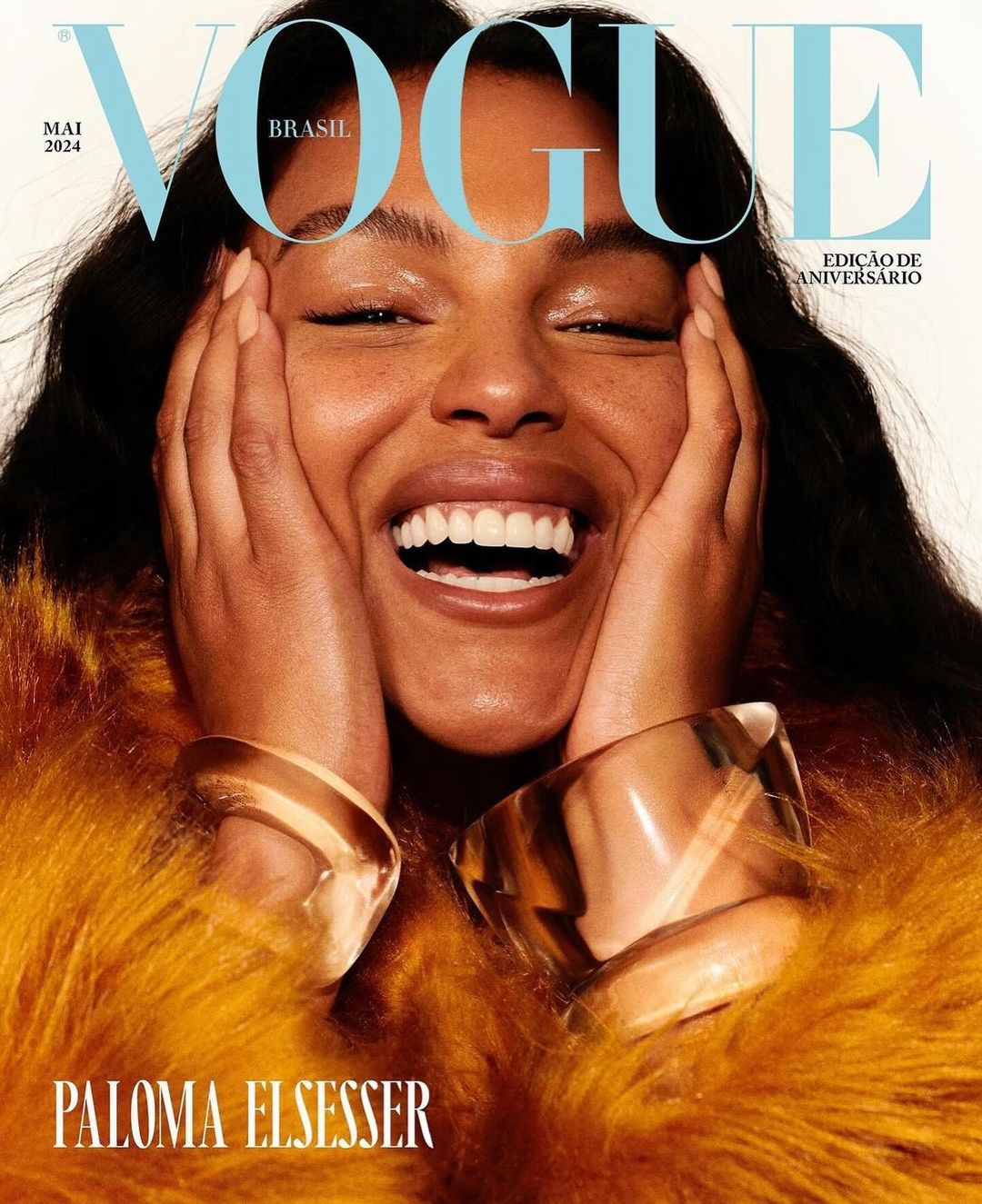 Vogue Brasil cover featuring Paloma Elsesser smiling widely, wearing gold bracelets and a yellow fur coat against a light background.