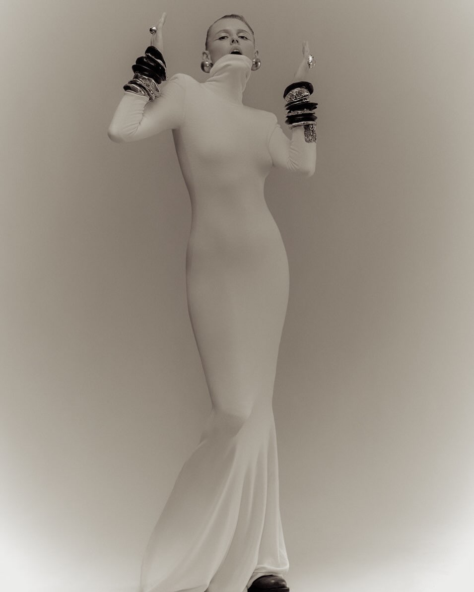 Woman in white dress and dark bangles strikes pose in front of neutral background. The portrait is clean and high fashion.