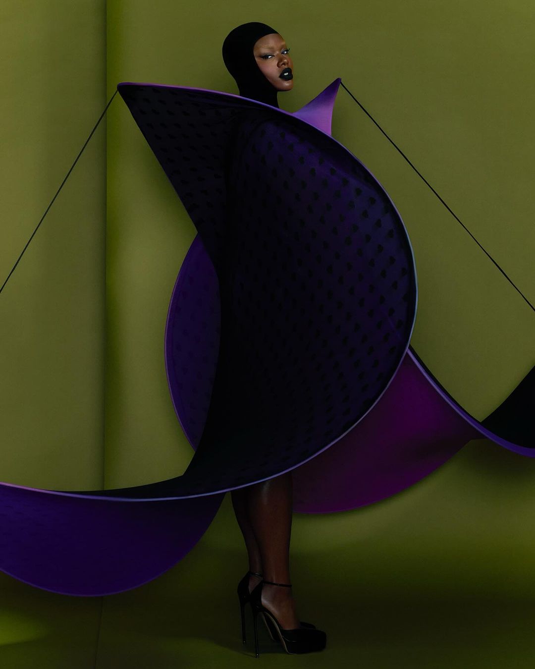 A fashion shot of a Black woman wearing a black hood and purple sculptural garment with black heels on a green background.