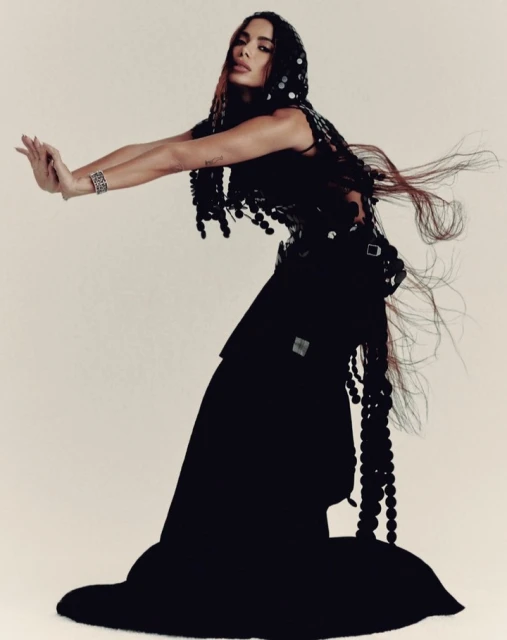 A woman with long, flowing hair and a black dress poses with her hands up against a plain background, looking at the camera.
