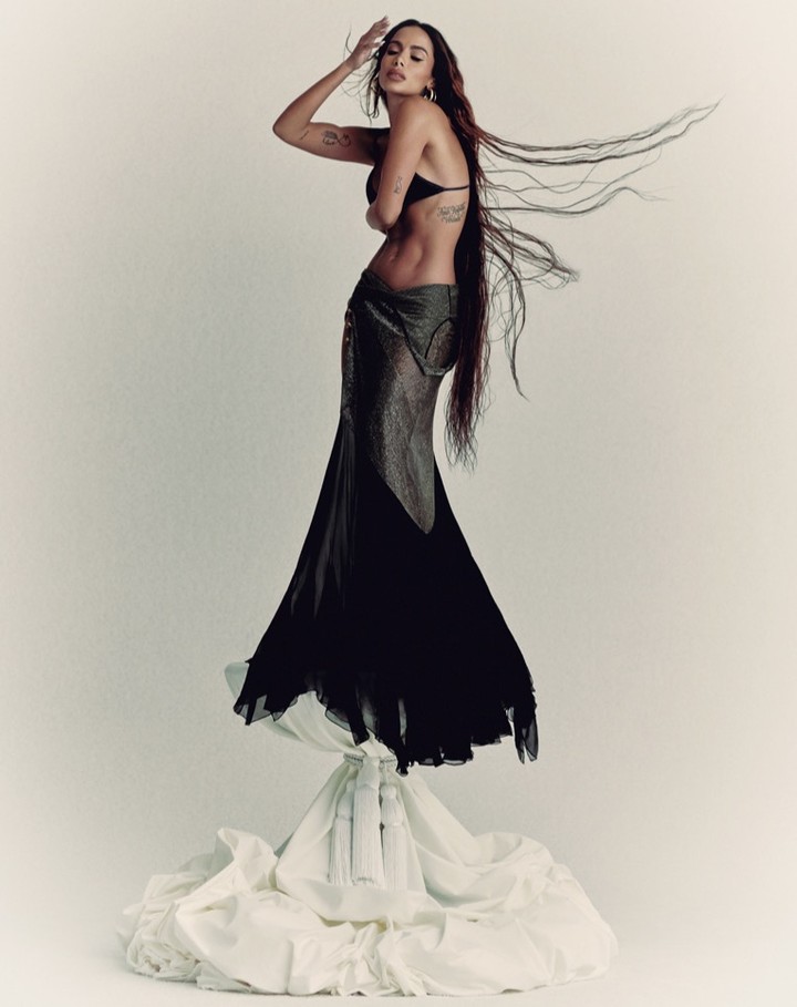 A woman with long brown hair in a black bra and flowing black skirt poses on a white, draped surface against a white background.
