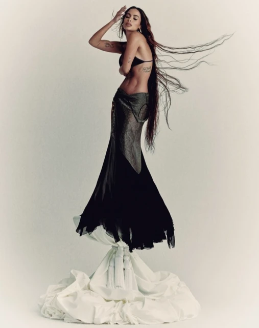 A woman with long brown hair in a black bra and flowing black skirt poses on a white, draped surface against a white background.