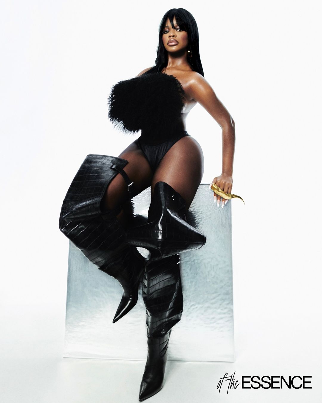 A confident Black woman in black one-piece and thigh-high crocodile-texture boots poses on a clear cube in a high-key studio shot.