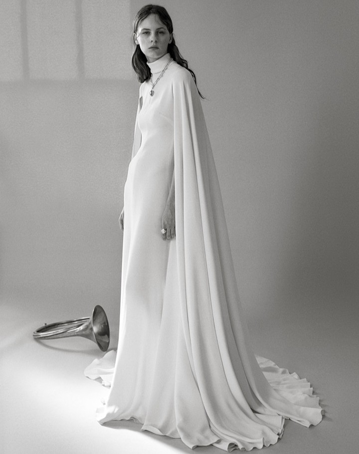 Black-and-white portrait of a woman in a white dress with cape detail, standing next to a flugelhorn in a bright studio setting.