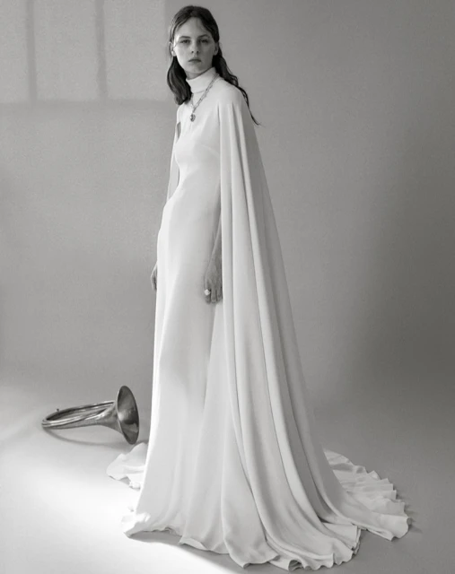 Black-and-white portrait of a woman in a white dress with cape detail, standing next to a flugelhorn in a bright studio setting.