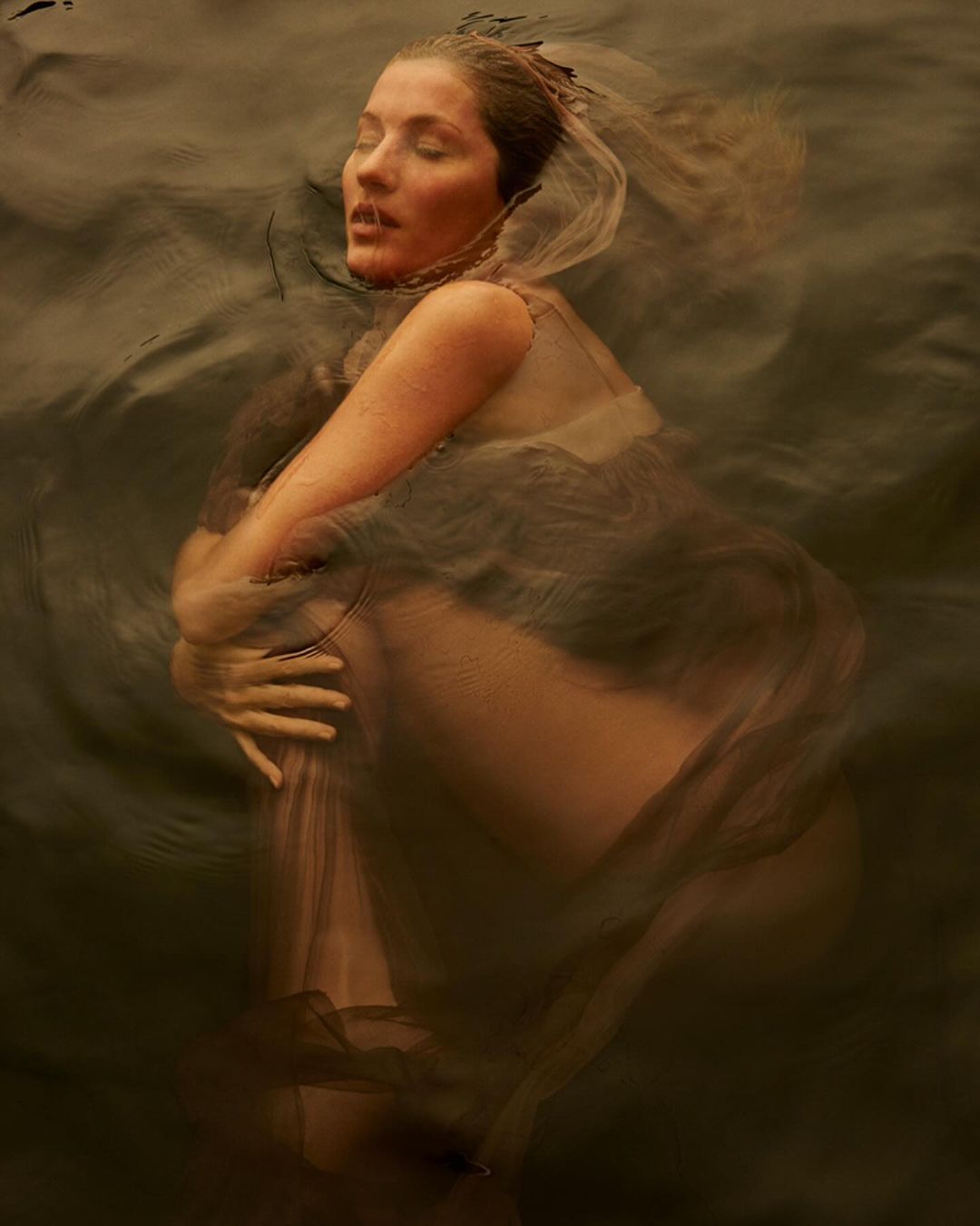 Woman with eyes closed embraces herself while submerged in water, sheer veil around her head, creating a serene and artistic underwater portrait.