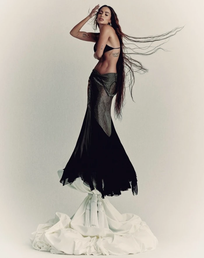 Woman with long dark hair blowing in the wind wearing a black top and long dark skirt standing on draped white fabric against a white seamless backdrop