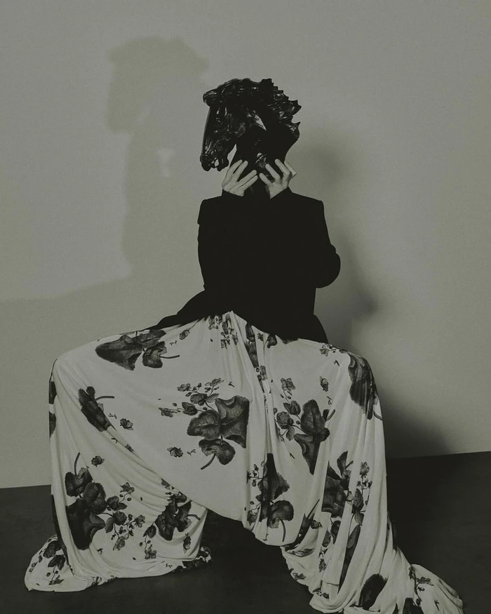 Model seated wearing a black jacket and large white floral patterned skirt while holding a black horse head mask over their face