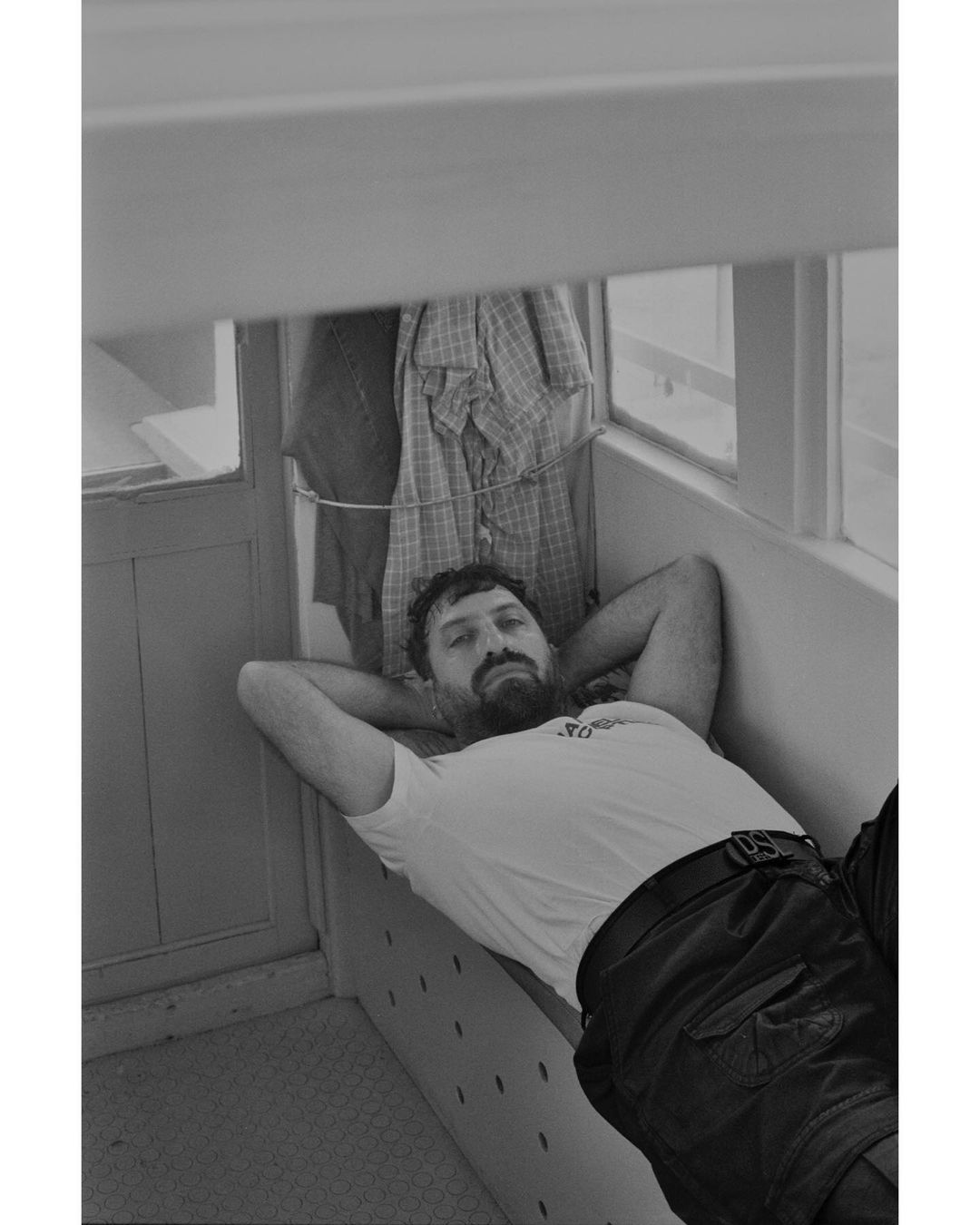 Man wearing a white t-shirt and dark trousers resting with his hands behind his head inside an interior cabin space.