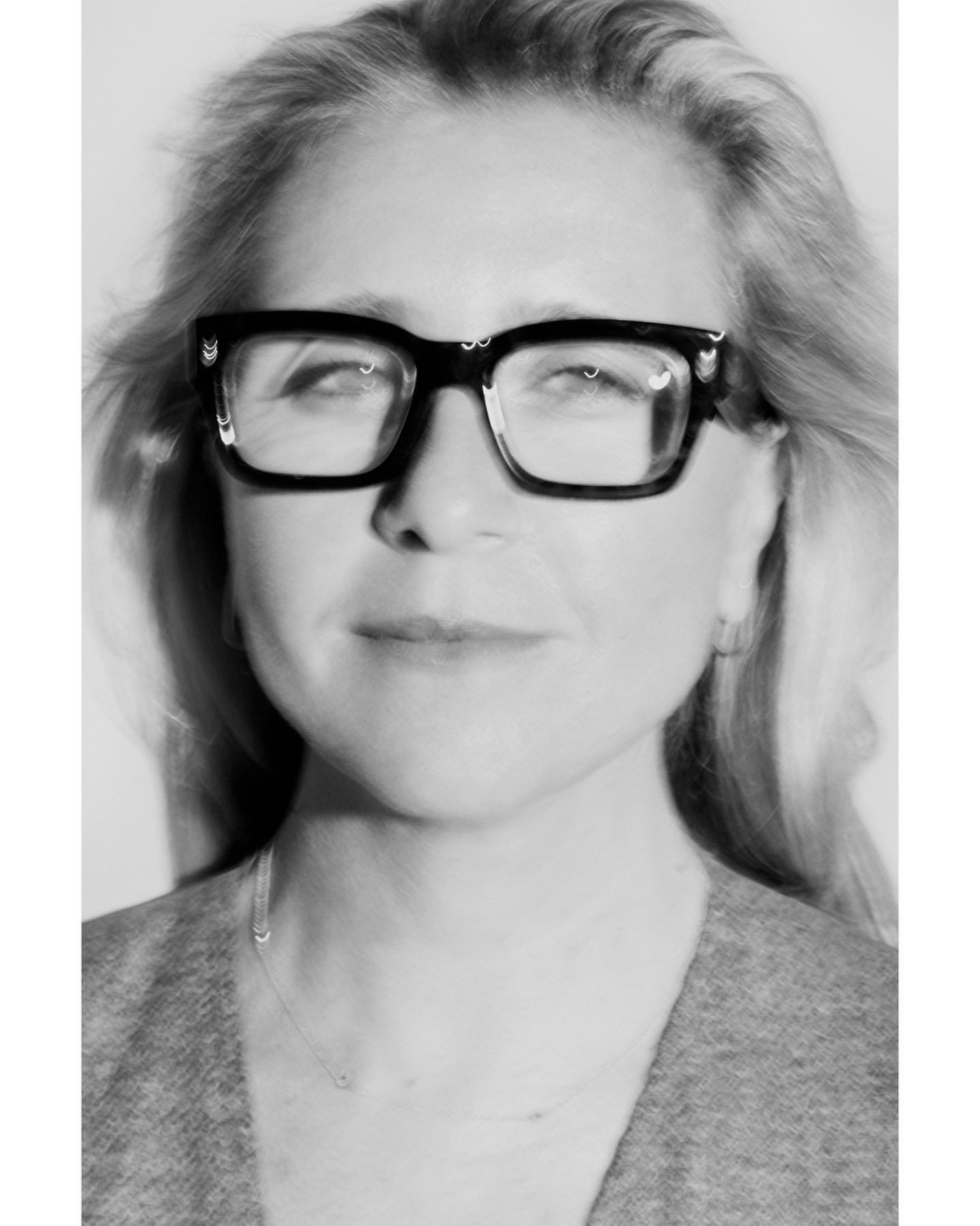 Black and white portrait of a mature woman wearing black-rimmed glasses and a gray sweater, captured in a well-lit indoor environment.