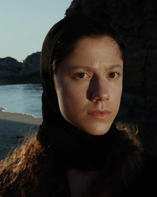 Close-up portrait of a woman with curly hair wearing a black headscarf, set against a backdrop of beach, rocks, and gentle waves.