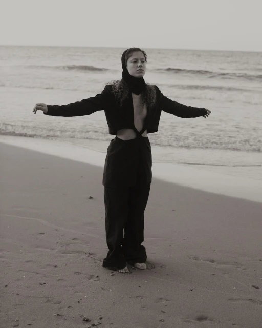 Monochrome outdoor portrait of a woman on a beach with arms outstretched, wearing a cropped jacket and loose pants.